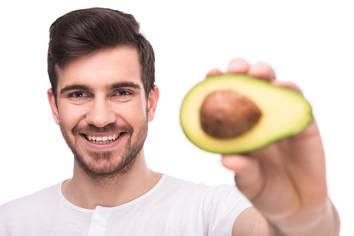 Handsome young man is holding avocado, standing against white background and smiling.Man is holding avocado, isolated on white background.