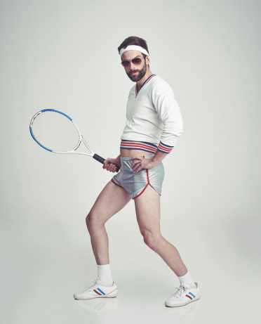 A young man in the studio wearing ill-fitting retro tennis wear and shades while holding a racquet