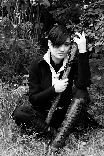 Vertical B&W outdoor garden shot of young Asian man, front view, sitting and smiling, holding katana in both hands.