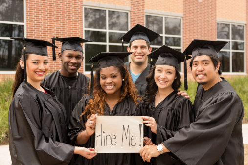 Six multi-ethnic graduates hold 'Hire Me' sign after college graduation. School building background. Employment issues concept.