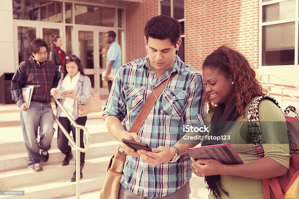 Education: Multi-ethnic group of college students talk, study before class. Multi-ethnic group of college studens studying and talking outside school building before class begins.  Latin male, African descent female listen to music on digital tablet in foreground. Cross processed. 20-24 Years Stock Photo