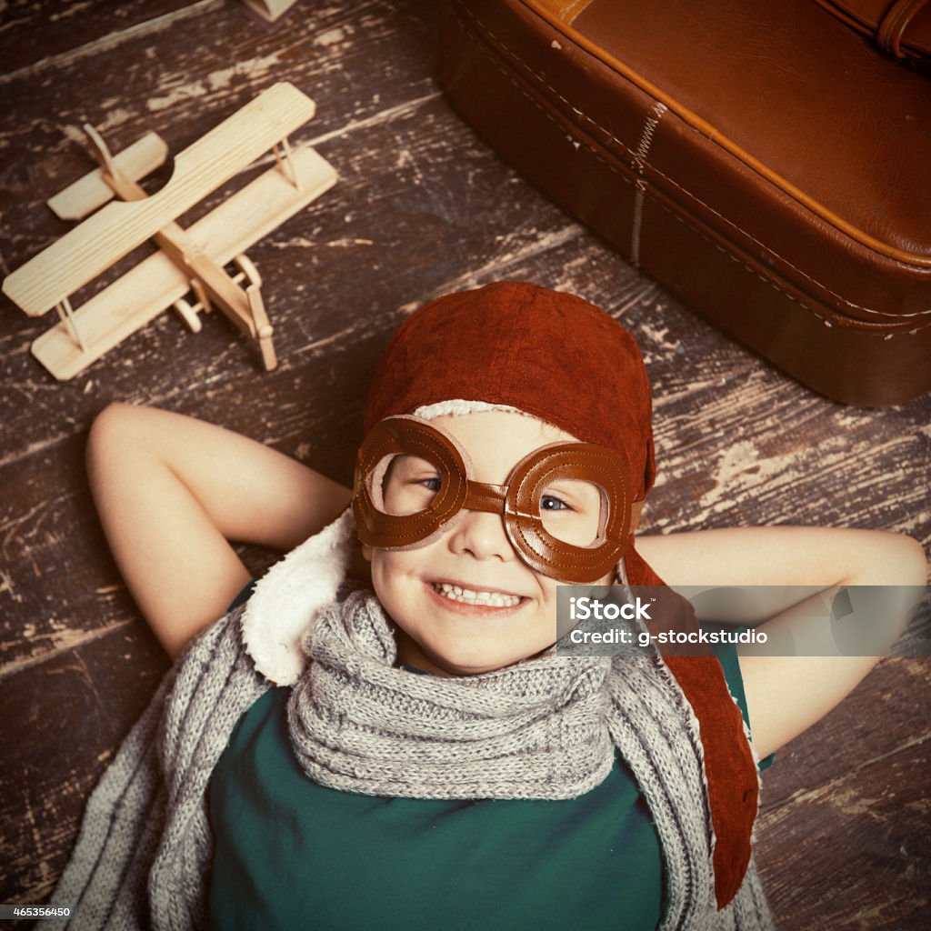 Happy little dreamer. Top view of happy little boy in pilot headwear and eyeglasses lying on the hardwood floor and smiling while wooden planer and briefcase laying near him 2015 Stock Photo