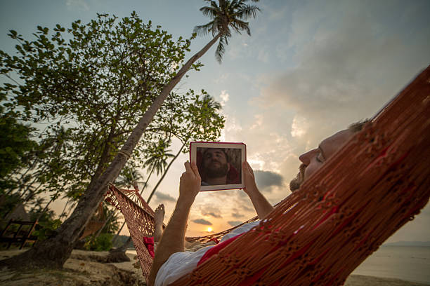 Man resting on hammock on tropical beach-Using digital tablet Young man lying down on hammock relaxing and using a digital tablet. Sunset on Koh Mak Island, Thailand. hammock men lying down digital tablet stock pictures, royalty-free photos & images