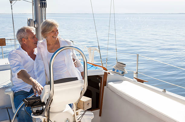 Happy Senior Couple Sailing Yacht or Sail Boat A happy senior couple sailing and sitting at the wheel of a sail boat on a calm blue sea old boat stock pictures, royalty-free photos & images