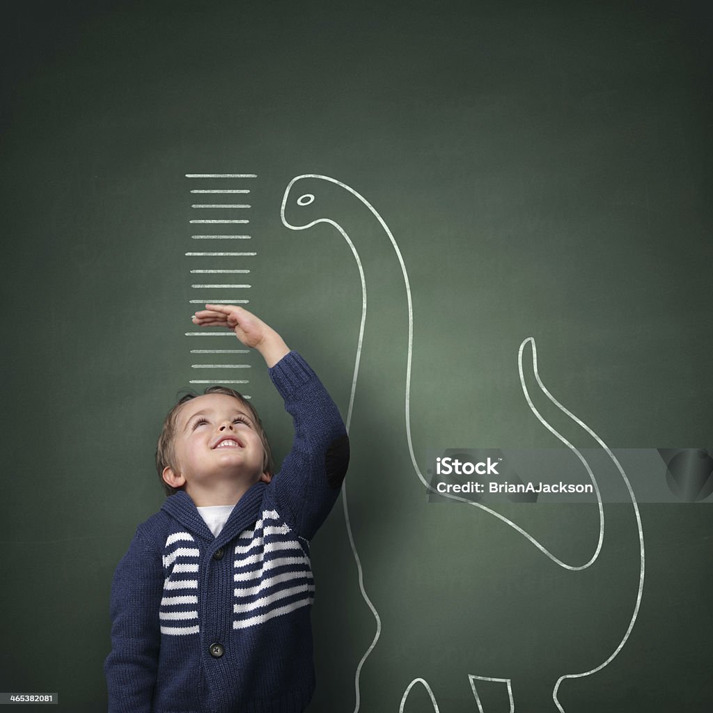 Growing up taller than a dinosaur Young boy measuring his growth in height against a blackboard with chalk dinosaur scale Child Stock Photo