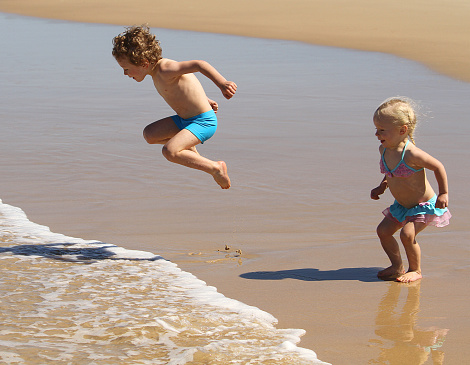 A little boy and girl, brother & sister, jumping waves at the beach together, wearing swimmers, having fun.