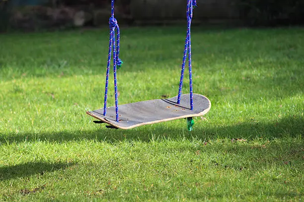 Photo showing a garden swing that has been made from an old skateboard, as part of a recycling project at school.  The upcycled skateboard swing has been tied to the branch of an apple tree with blue rope, suspending it above the lawn.