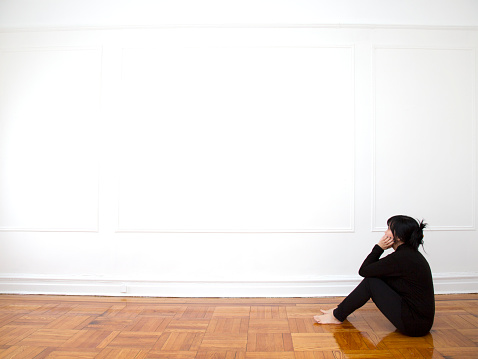 Asian woman in all black clothing sits with her elbows on her knees on her wooden, parquet floors in her empty bedroom in her apartment looking up at a blank white wall. Photo:  T. Tan   2015 © Tan.  All Rights Reserved.