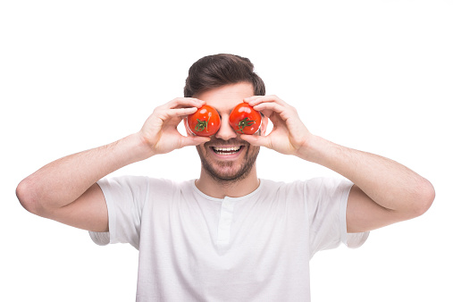 Handsome young man is holding tomatoes in front of his eyes while standing against white background.