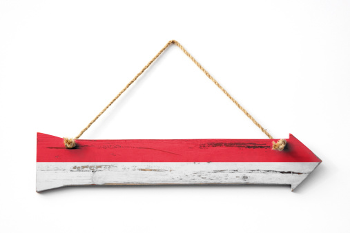 A wooden arrow, hanging against a white wall via a rustic rope, with the Indonesian flag stenciled upon it.