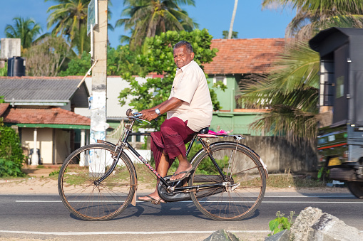 Hikkaduwa, Sri Lanka - February 24, 2014: Elderly man in sarong and shirt riding a bicycle. Cycling is the main transportation for the traditional people in the country.