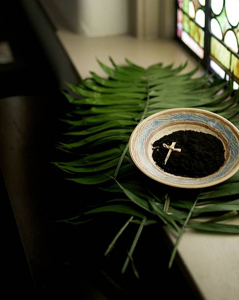 A cross in a bowl of dirt on top of leaves next to a window We prepare for the Lenten Season with the imposition of Ashes. The palm branches from last year during Holy Week are burned to prepare for this year’s Ash Wednesday. liturgy photos stock pictures, royalty-free photos & images