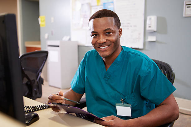 Portrait Of Male Nurse Working At Nurses Station Portrait Of Male Nurse Working At Nurses Station Smiling At Camera medical scrubs stock pictures, royalty-free photos & images