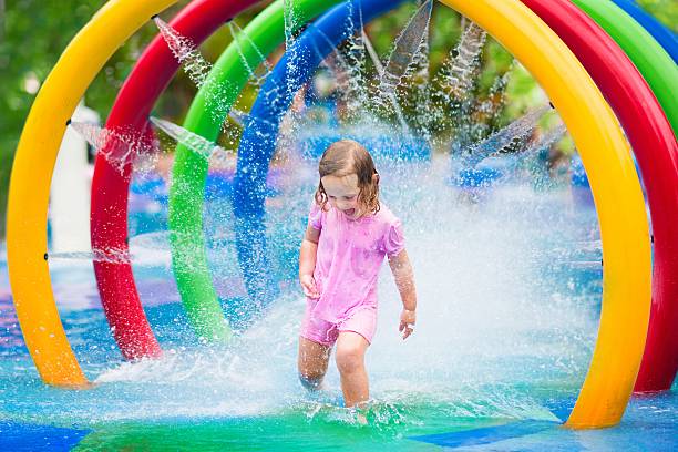 Happy little toddler girl running through a fountain having fun with water splashes in a swimming pool enjoying day trip to an aqua amusement park during summer family vacation