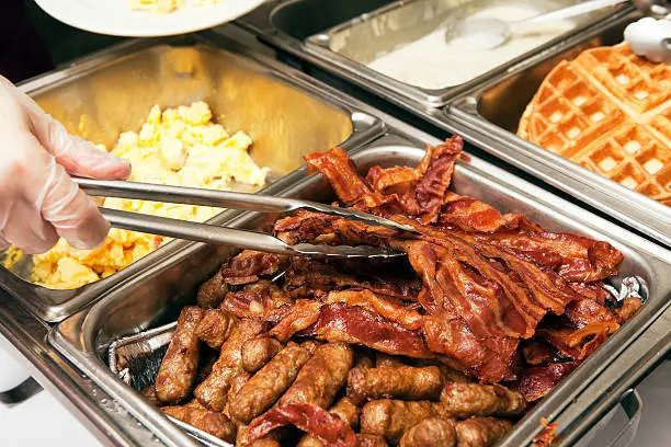 A gloved food service worker is  using tongs to plate bacon from a breakfast or brunch buffet line. This chafing dish has sausage, bacon and scrambled eggs, to the right are waffles and gravy for biscuits and gravy.