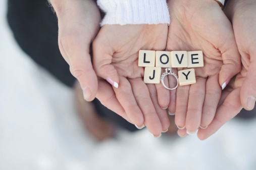 A closeup of a couple holding their engagement ring with the words love and joy.