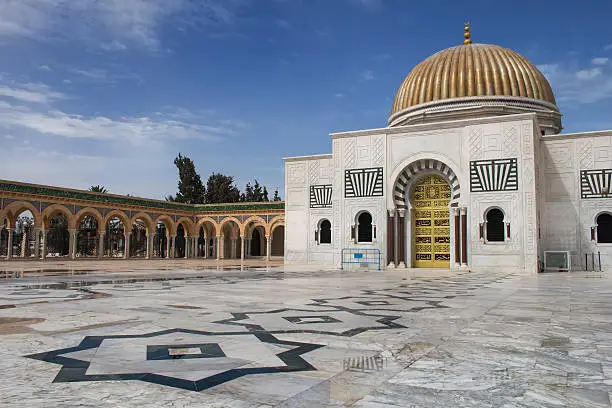White marble floor with black stars and white building of the Mausoleum of Habib Bourguiba in Monastir, Tunisia. Shiny golden dome, contrasting with the bright blue sky. Colonnade and a park. 