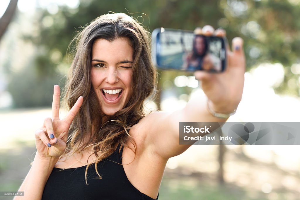 Young woman taking selfie in park with phone Portrait of a beautiful young woman selfie in the park with a smartphone doing v sign 2015 Stock Photo