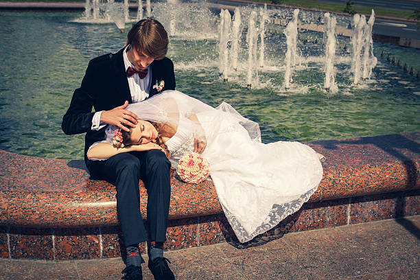 Bride in a white dress is lying by a water source, splashing water and holding her bouquet