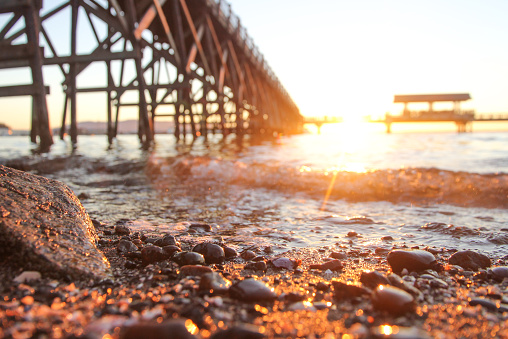 A DSLR, shallow depth of field photo of a rocky beach during golden hour. A dock and boardwalk sits blurred out in the background. Large and small rocks and pebbles line the short of the beach and the ocean water. The sun glows off the rocks and water. Located on the bay in bellingham, Washington