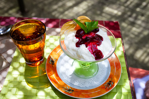 Spring time dessert at an outdoor terrace restaurant in Provence, France.  The coup glass contains ice cream and/or yoguhrt with red cherries, mint leaves and a kumquat.  There is also a glass of sparkling mineral water with bubbles.  The dessert sits on a soft green woven placemat, which rests on the ceramic tiled table top.  The dessert plate is decorated with an orange band and what appears to be olives on the branches of an olive tree.