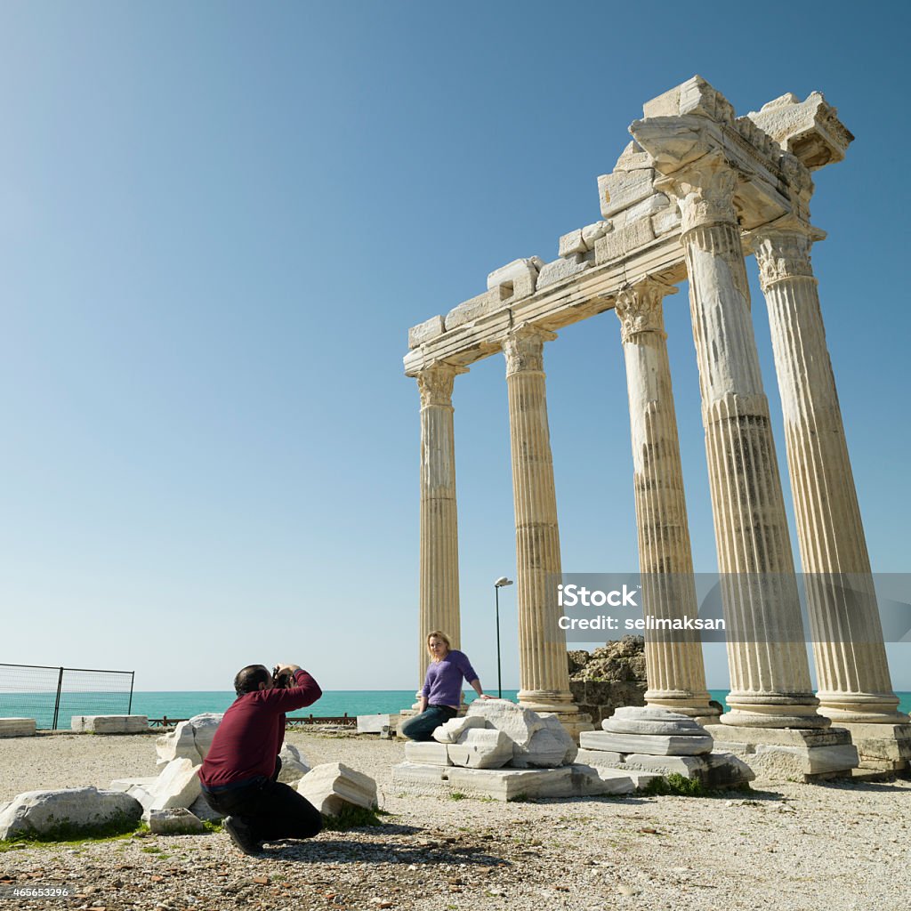 Young traveling couple taking photos in old ruins of Side Young traveling couple taking photos in old ruins of Apollo Temple in city of Side,Antalya,Turkey.The woman is standing next to columns of monument while man is taking photos via camera.Hasseblad H4D is used. Candid Stock Photo