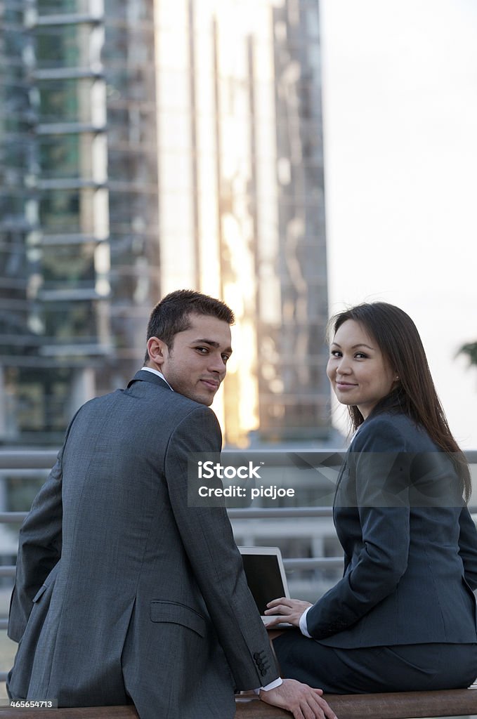 businessman and woman working on laptop in financial district businessman and businesswoman sitting on bench working on laptop in financial district looking at the camera 25-29 Years Stock Photo