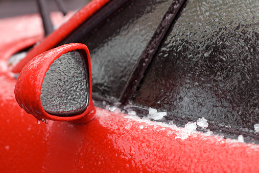Close up of ice coating a red car's mirror, door and windows during a winter storm.