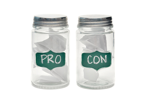 Closeup of a jars labelled Pro and Con with pieces of paper in them on a white background with shadows.