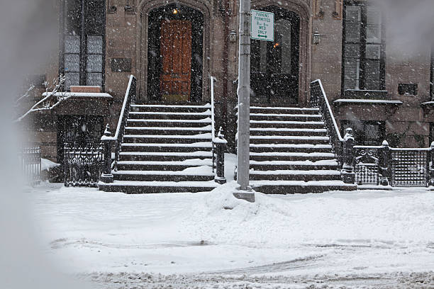 Snow covered Brooklyn brownstone townhouse stoops and sidewalk in winter. Snow is falling during a late winter storm in the Brooklyn Heights Historic District. Gardens, stoops, and sidewalks are covered in deep snow. The snow plow has not yet cleared the street.