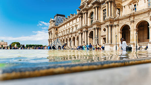 Paris, France - August 18, 2014: Tourists visitig Louvre - Cour Napoleon in summer with relaxed people sitting on the edge of fountain near the Pyramid by the architect Ieoh Ming Pei. Louvre is the most visited museum in the world. Paris sightseeing