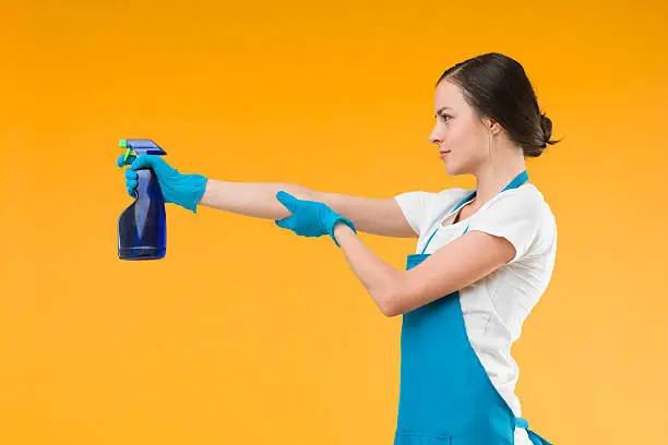 side view of cleaning woman pointing spray with liquid detergent, shooting, against yellow background