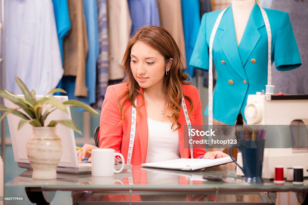 Latin descent, woman small business owner. Tailor, fashion designer, boutique. Latin descent, female small business owner at work. She could be a tailor doing alterations, a fashion designer creating new clothing trends, or a boutique owner.  She is using her laptop computer to place an online fabric order at her desk. Sewing machine to right, clothing hanging on racks and a mannequin with tape measure in background. Minority Groups Stock Photo