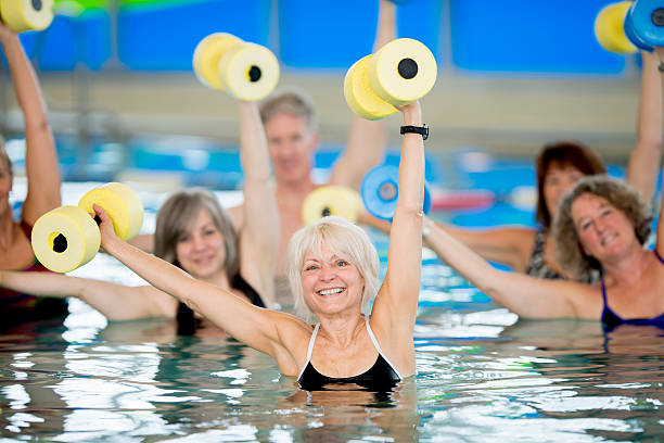 Group of Seniors doing Water Aerobics A group of seniors practicing water aerobics in an indoor pool, holding dumbbells over their heads. aquatic sport stock pictures, royalty-free photos & images