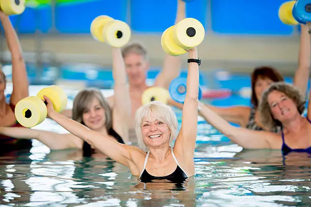 A group of seniors practicing water aerobics in an indoor pool, holding dumbbells over their heads.