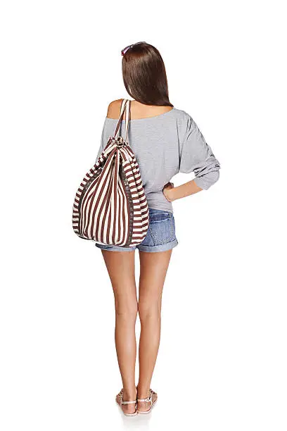 Back view Full length of young slim tanned female in denim shorts with backpack, isolated on white background
