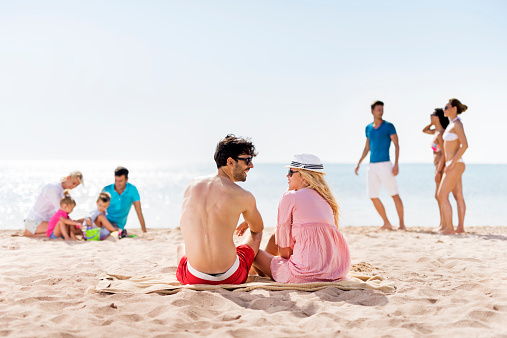 Group of people enjoying a day on the beach. Focus is on foreground, on young couple sitting and talking.