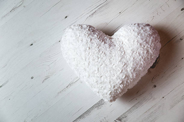 Fluffy soft decorative white heart shaped pillow on bleached floorboard surface as viewed from above. Sidelight studio soft flash with deep shadow. Shot on Canon EOS. Edie with soft muted blacks and minor grain.