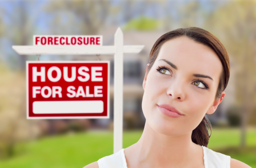 Thoughtful Pretty Mixed Race Woman In Front of Home and Foreclosure House For Sale Real Estate Sign Looking Up and to the Side.