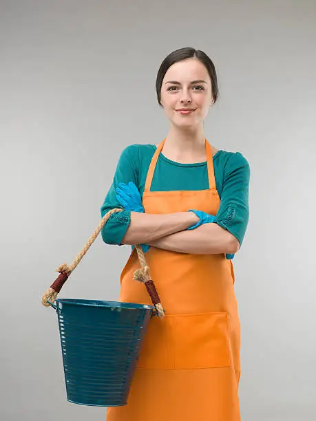 happy confident cleaning lady holding bucket with arms crossed, on grey background