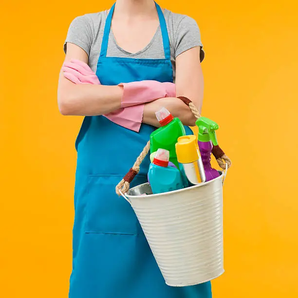 woman in blue apron holding metal bucket with cleaning supplies against yellow background