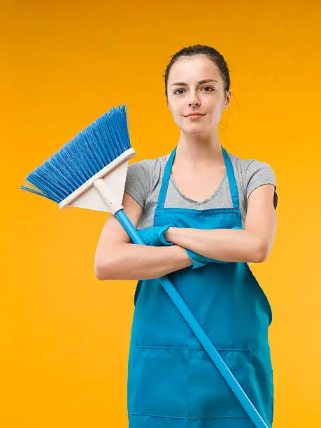 happy caucasian woman standing with arms crossed and holding a broom, on yellow background