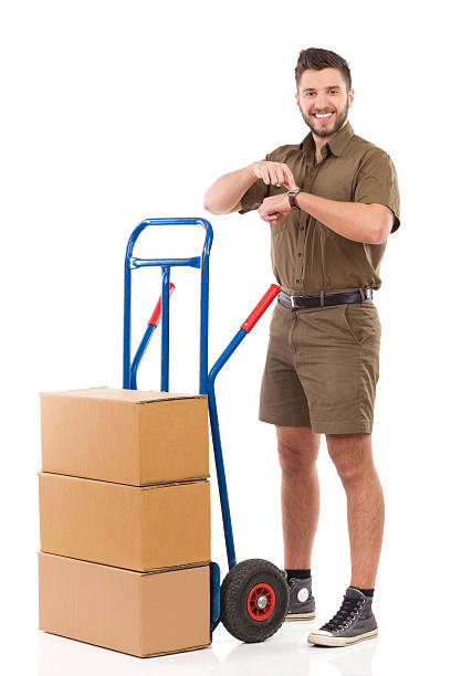Just in time Delivery man standing with a push cart and pointing at wristwatch. Full length studio shot isolated on white. mover delivery person box hand truck stock pictures, royalty-free photos & images