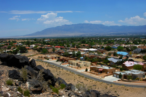 Overlook at the Petroglyph National Monument shows Albuquerque, New Mexico.  Sandia Mountains loom in the background and in the foreground black basalt boulders and a curving city street.