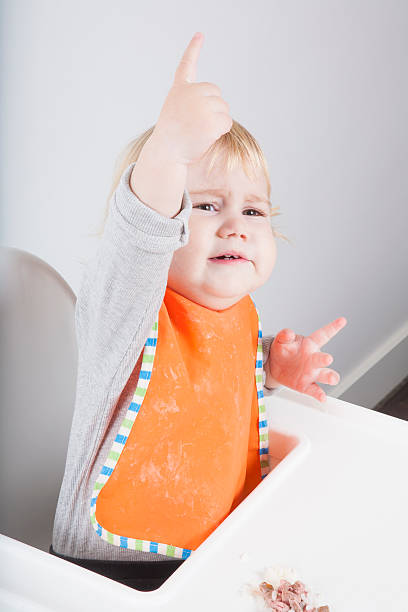 calling in lunch blonde caucasian baby seventeen month age orange bib grey sweater eating tuna rice on white high-chair asking with hand up kids eating tuna stock pictures, royalty-free photos & images