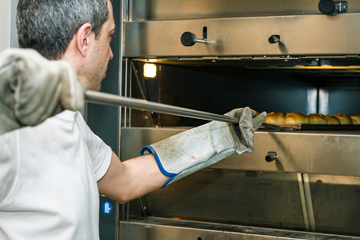 Adult man (baker) inserting breads in to oven using baker-shovel