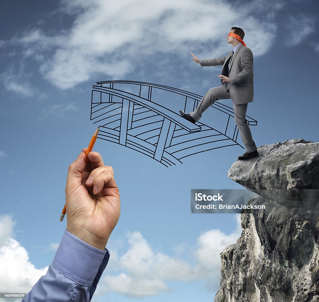 Building bridges - assistance for business Businessman in a blindfold stepping off a cliff ledge with giant hand drawing a bridge for a safe crossing concept for building bridges, risk, challenge, conquering adversity, ignorance and assistance Danger Stock Photo