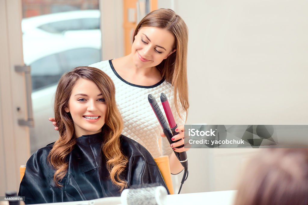 Beautiful woman in hair salon Going for big curls. Mirror reflection of a young beautiful woman discussing hairstyling with her hairdresser while sitting in the hair salon and getting her hair done with hair iron 2015 Stock Photo