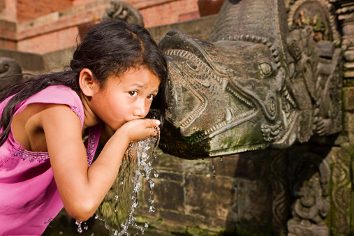 Young Nepali schoolgirl drinking from city fountain (founded by King Veer Deva in 299 A.D.) on Durbar Square Patan, Nepal. Patan Durbar Square has been listed by UNESCO as one of seven Monument Zones that make up the Kathmandu Valley World Heritage Site.