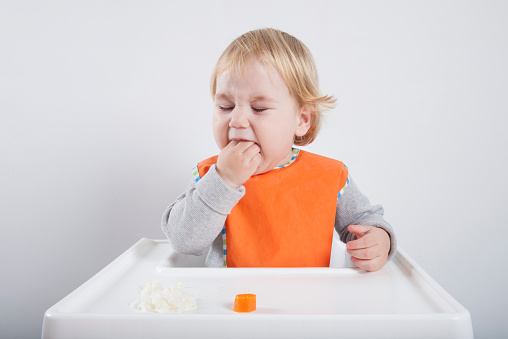 blonde caucasian baby seventeen month age orange bib grey sweater eating meal with her hand in white high-chair